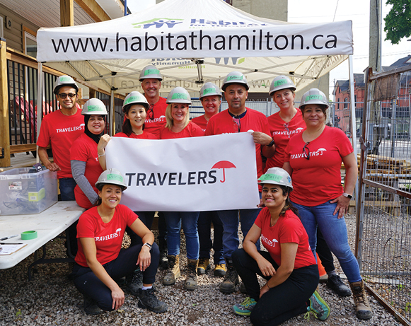 eleven people in red T-shirts and hardhats posing under a Habitat Hamilton tent with a Travelers banner