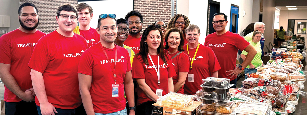 twelve people in Travelers T-shirts standing behind a long table laden with groceries