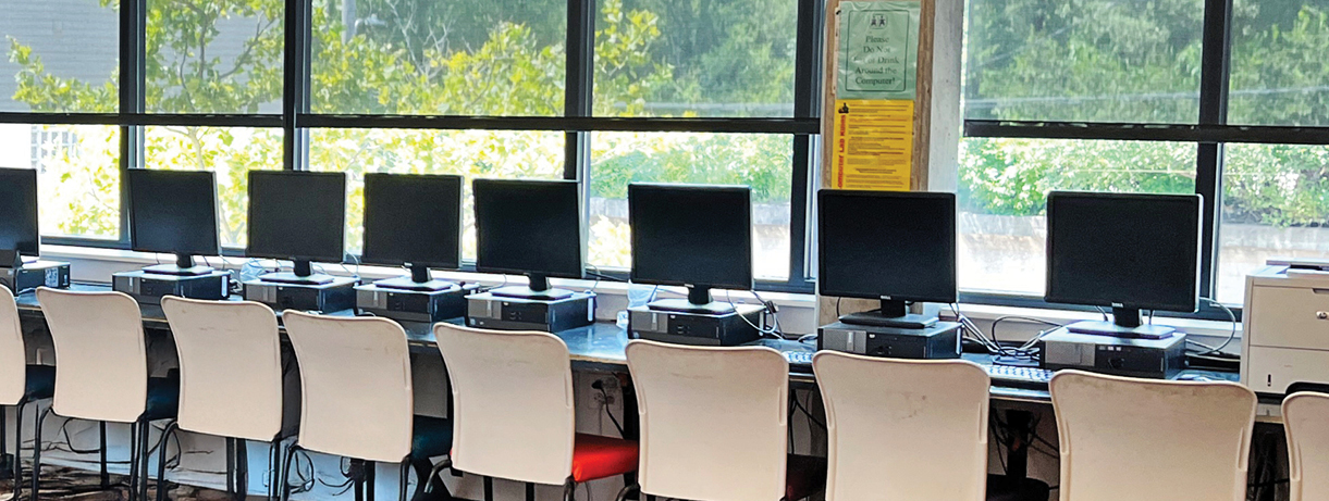 a row of computers on a long table by a window in a computer lab